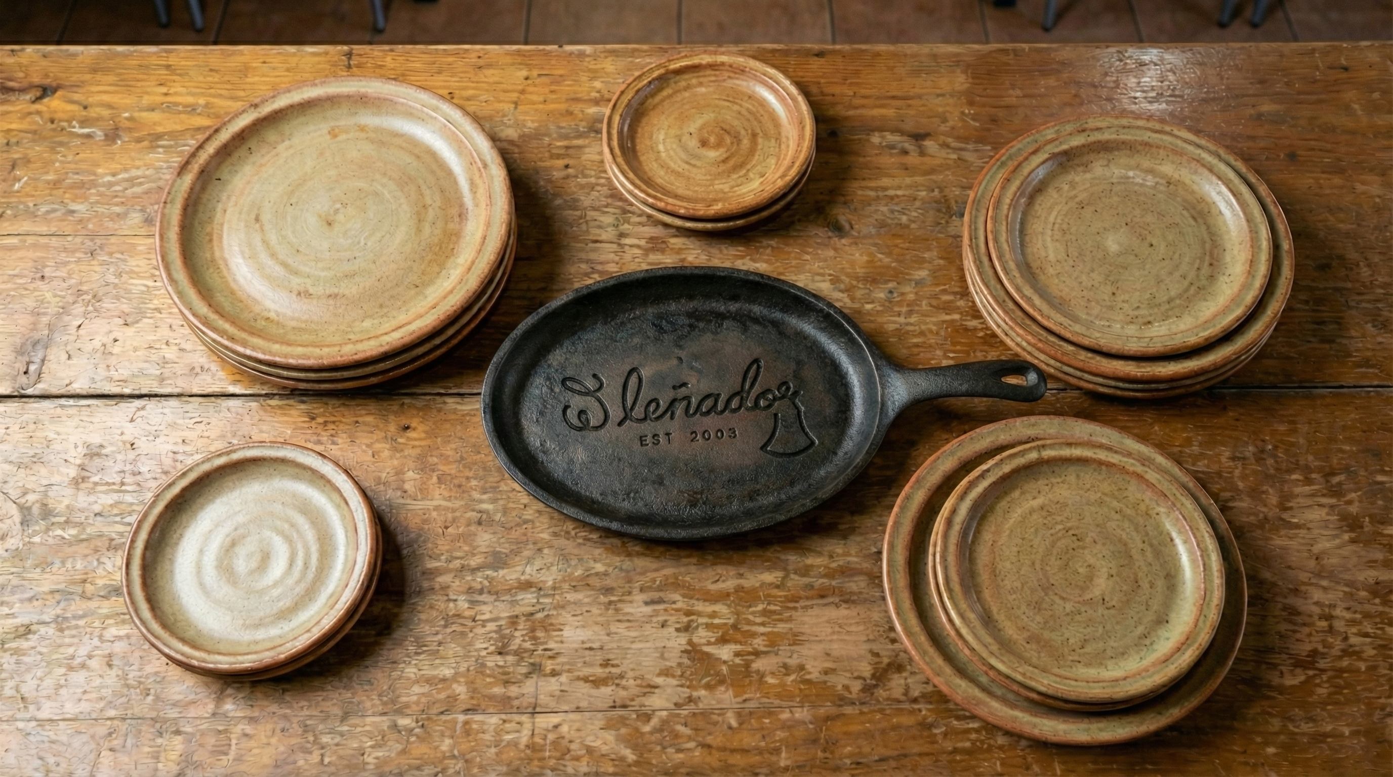 A rustic wooden table holds an assortment of earthy-toned plates and a cast-iron skillet with the 'El Leñador' logo embossed on.